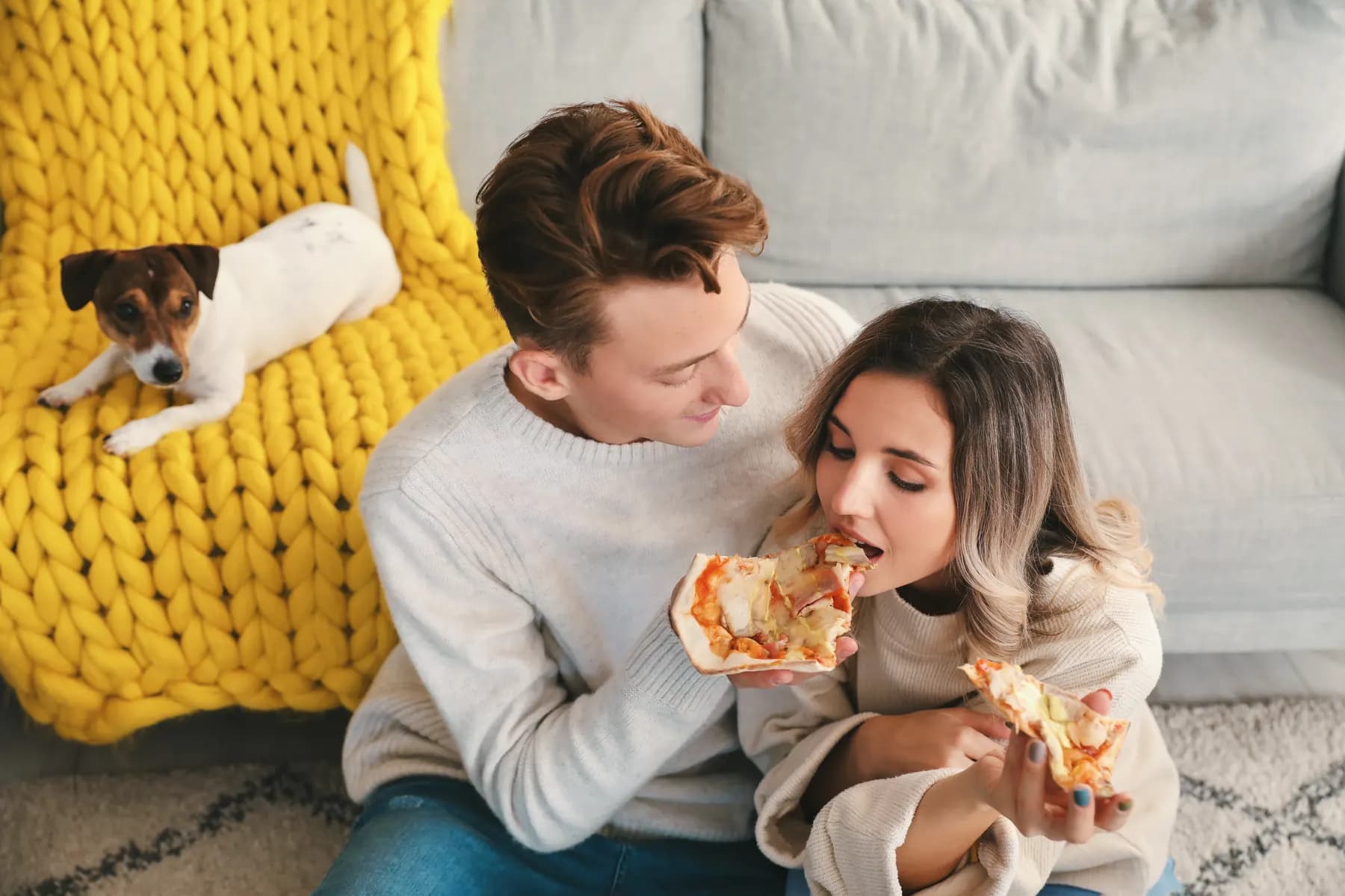 A Jack Russell terrier relaxing on a yellow knit blanket on the sofa while its owners share a pizza