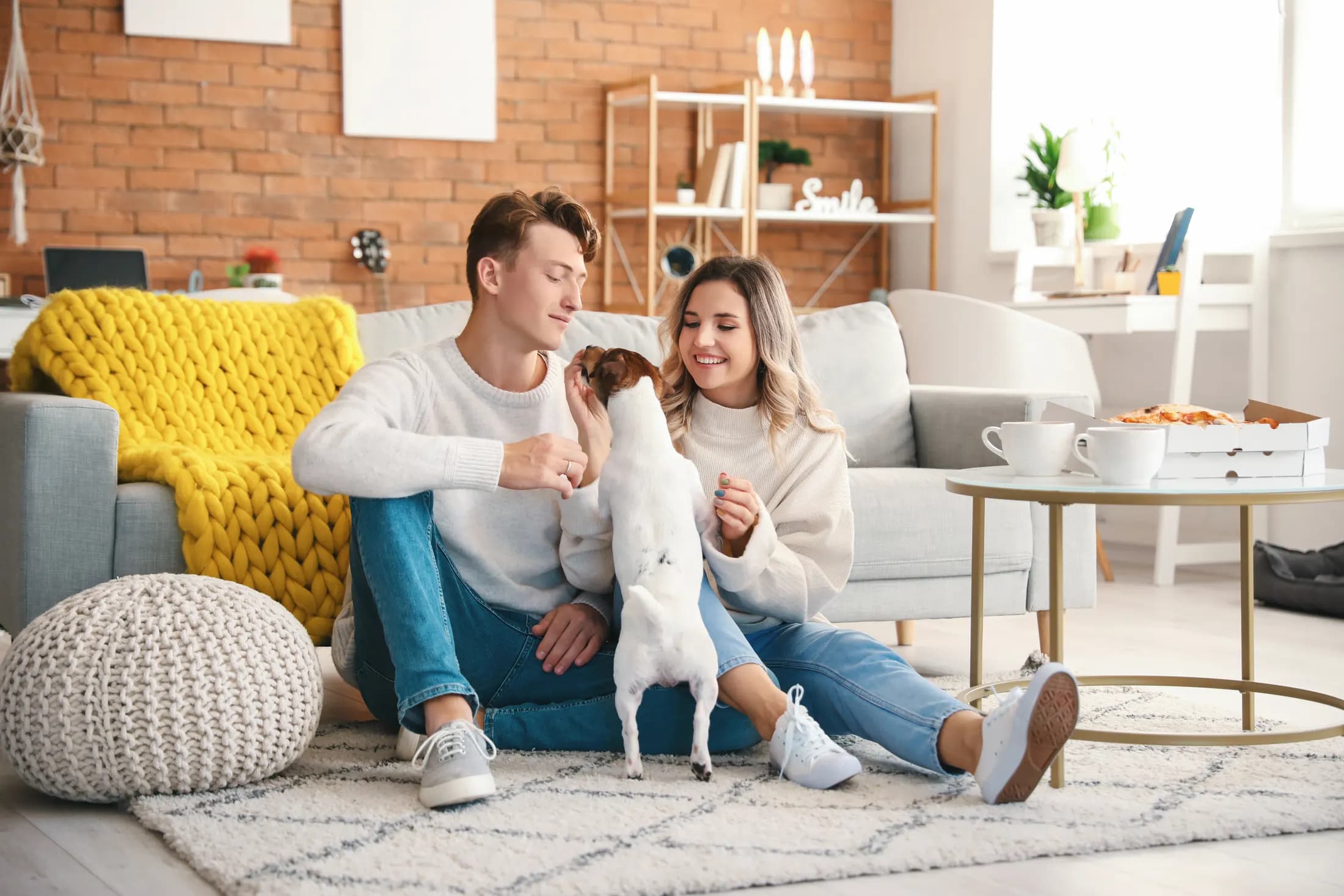 A couple at home on the living room rug, playing with their Jack Russell terrier next to a yellow knit blanket